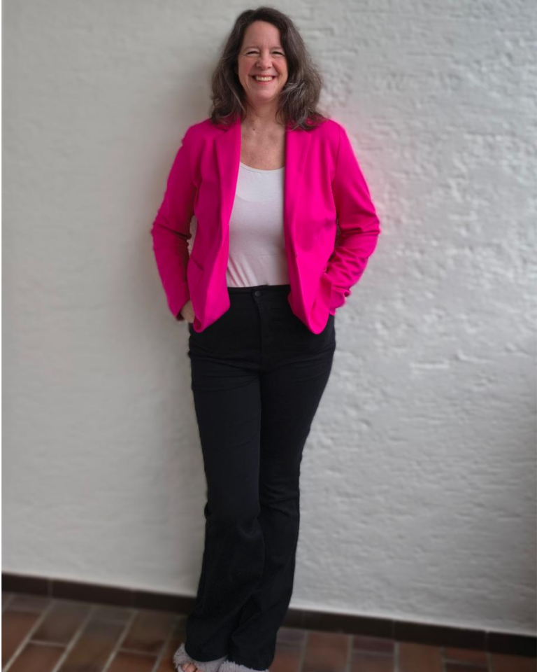 Charlotte steht in pink Sacko und schwarzer Jeans entspannt vor einer weißen Wand und dunklen Fließen